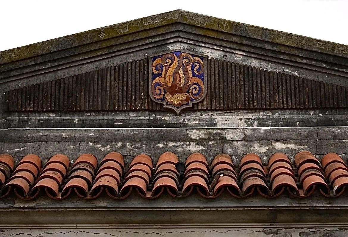 El frontis de una construcción antigua, con un monograma de mosaico sobre las tejas españolas.